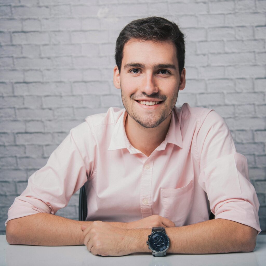 Confident young man in a pink shirt smiling at the camera against a brick wall backdrop.